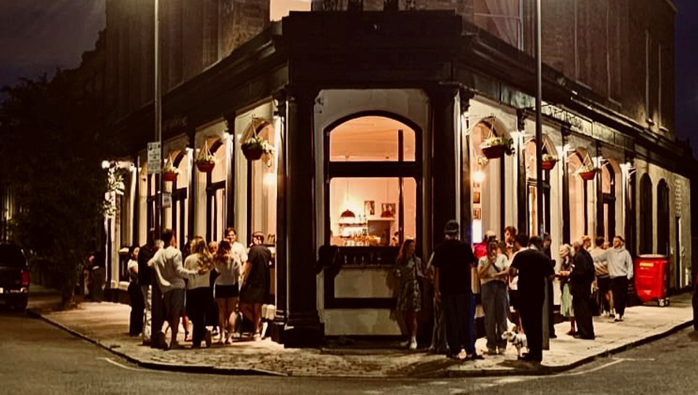 Exterior shot at night of the Queens Arms in Battersea, with revellers outside in front of big bay windows illuminated with a warm orange light