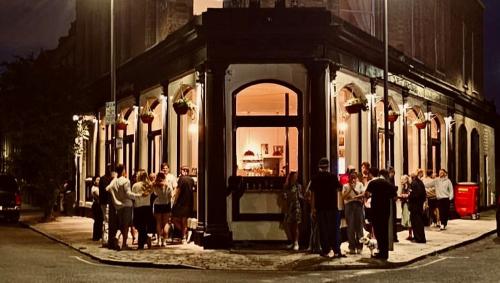 Exterior shot at night of the Queens Arms in Battersea, with revellers outside in front of big bay windows illuminated with a warm orange light