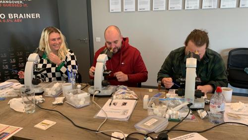 A female and two males sit at a desk with laboratory equipment in front of them including microscopes and chemical jars